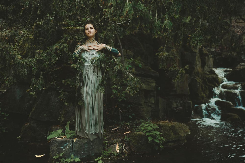 A person with painted arms and a long green dress stands beside a forest stream.