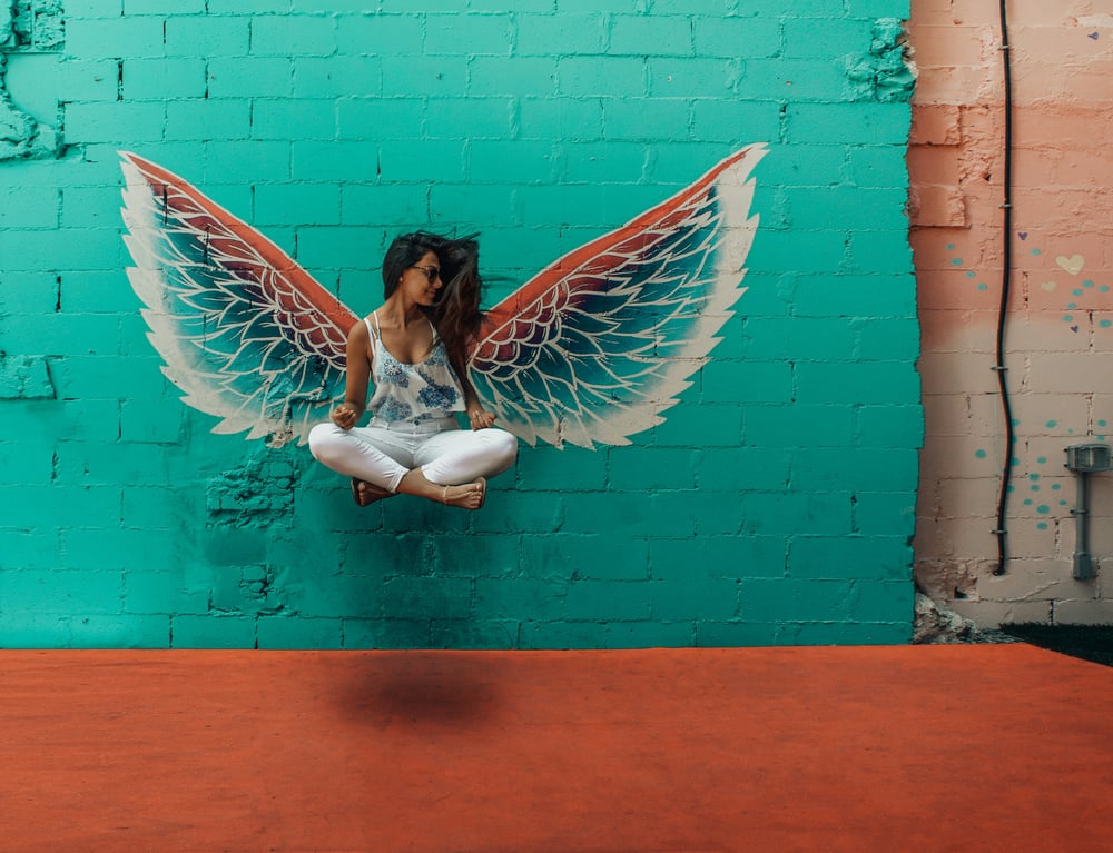 A person sits cross-legged and levitates in front of a wall with angel's wings painted on it.