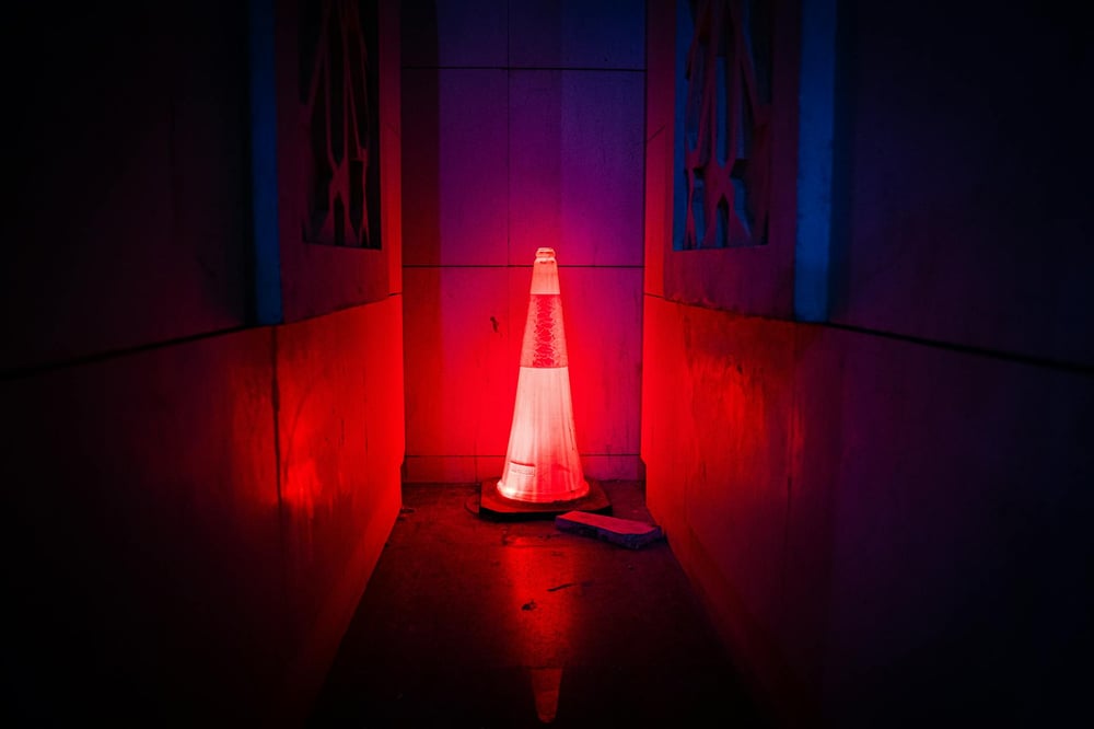A traffic cone is lit up by red light in a dark hallway.
