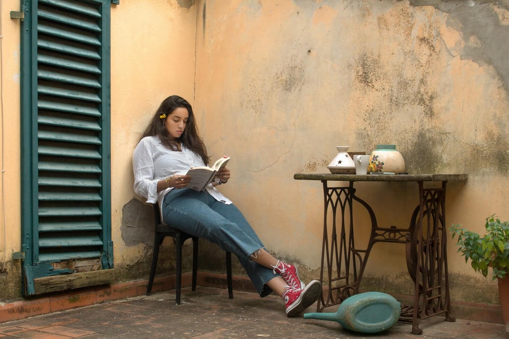 A person sits outside beside a worn yellow wall and reads a book.