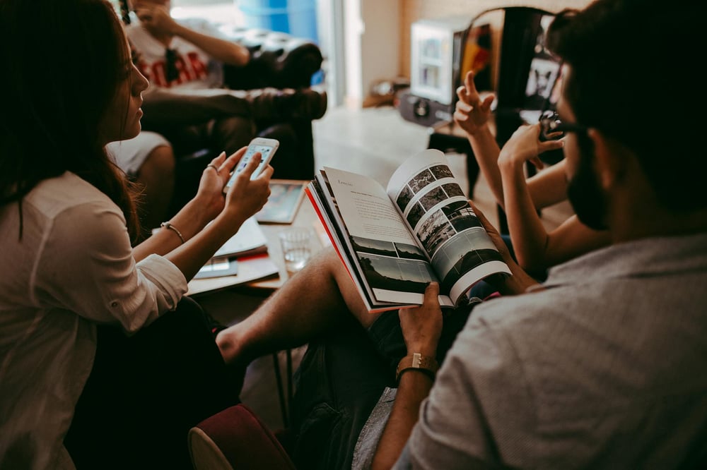 A small group of people gather around a coffee table, conversing over a book.