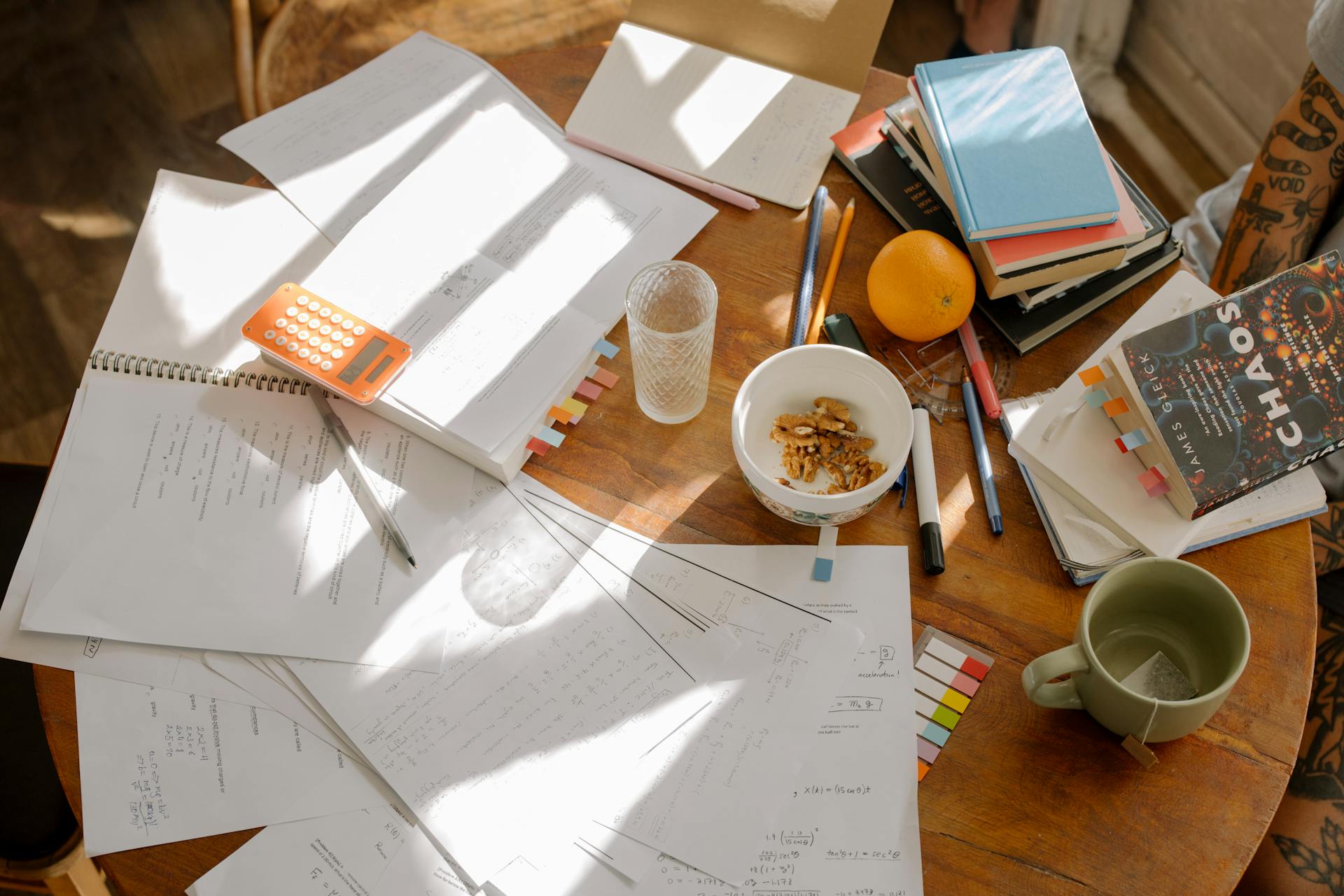 Ghostwriting kind of looks like this sometimes. A round table covered in a mess of notebooks, loose pages, stacked books with post-its sticking out of them, a bowl of pecans, an orange, a water glass, coffee cup, and a bunch of pens.