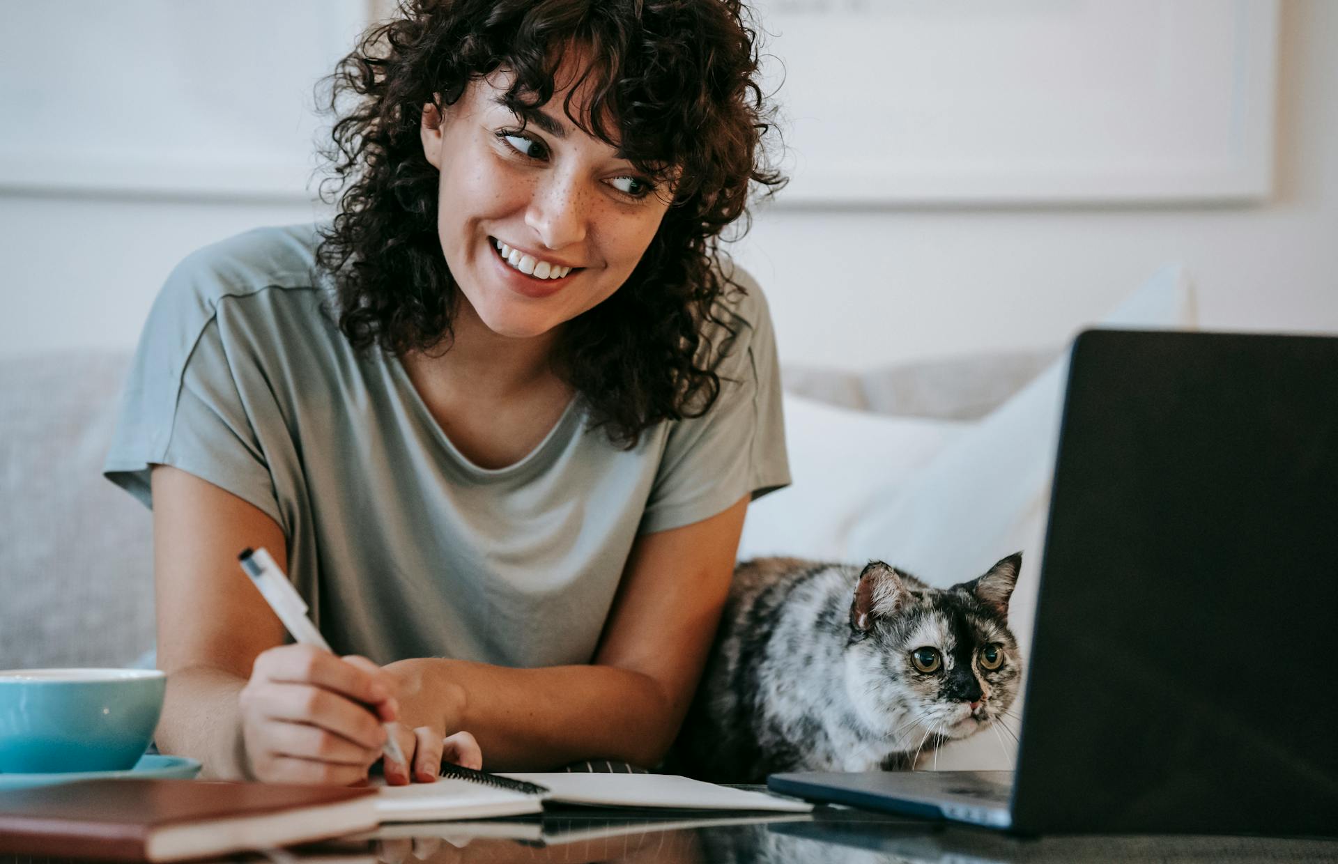 A person sits beside a cat and writes in a notebook while smiling at a computer screen.