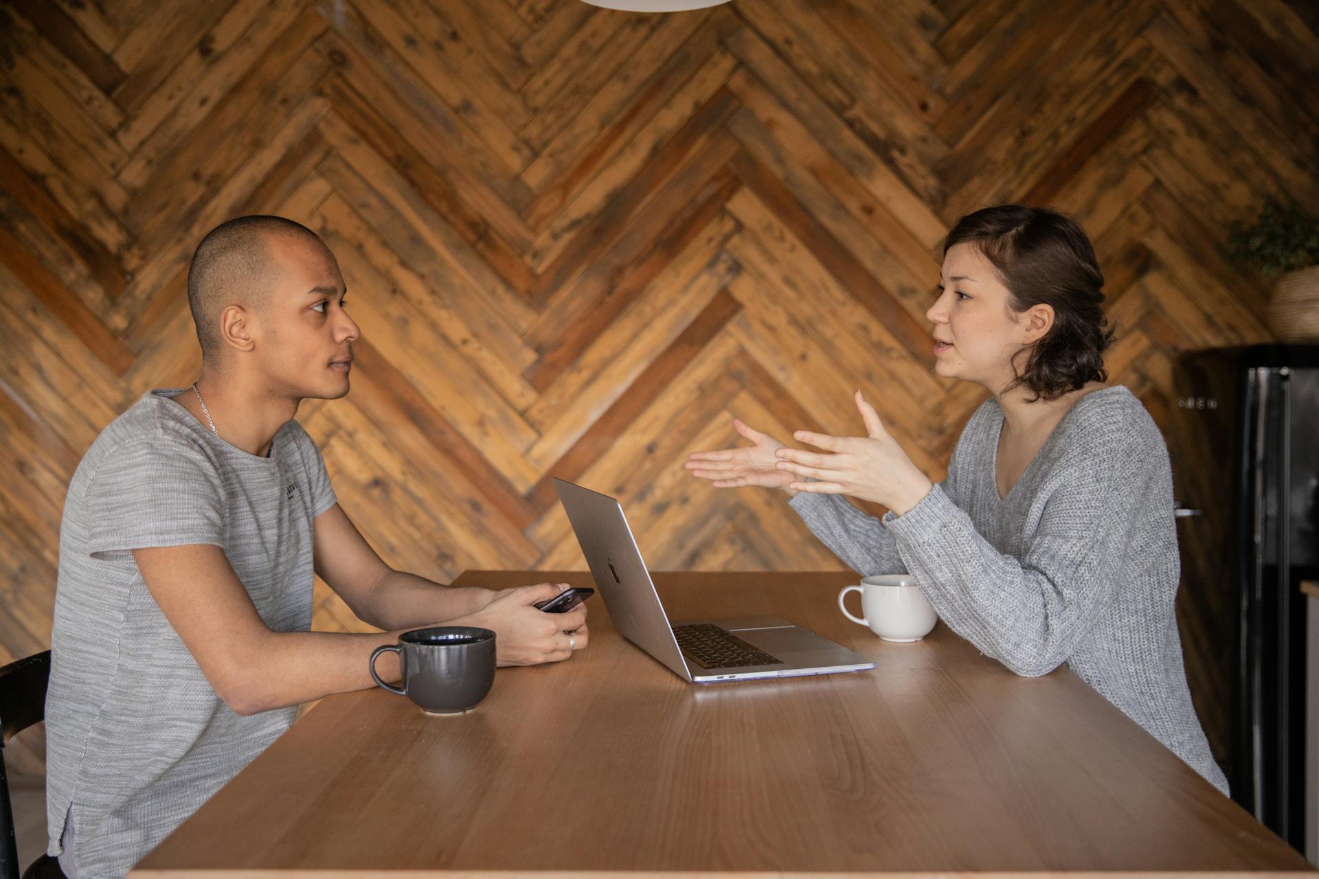 Two people sit on either side of a wooden table having a conversation with a laptop between them.