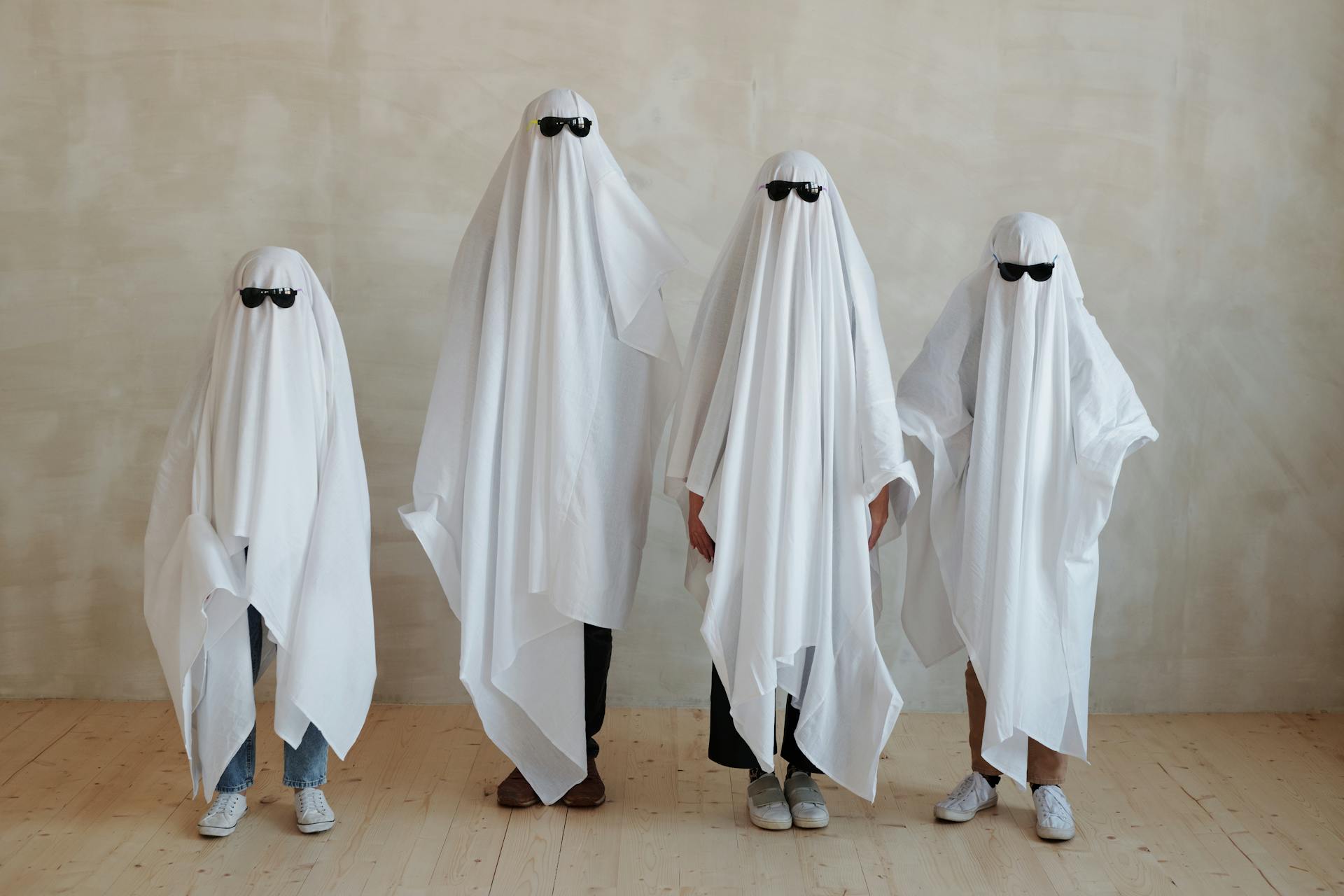 Four kids stand in a row wearing sheets like ghosts and sunglasses.