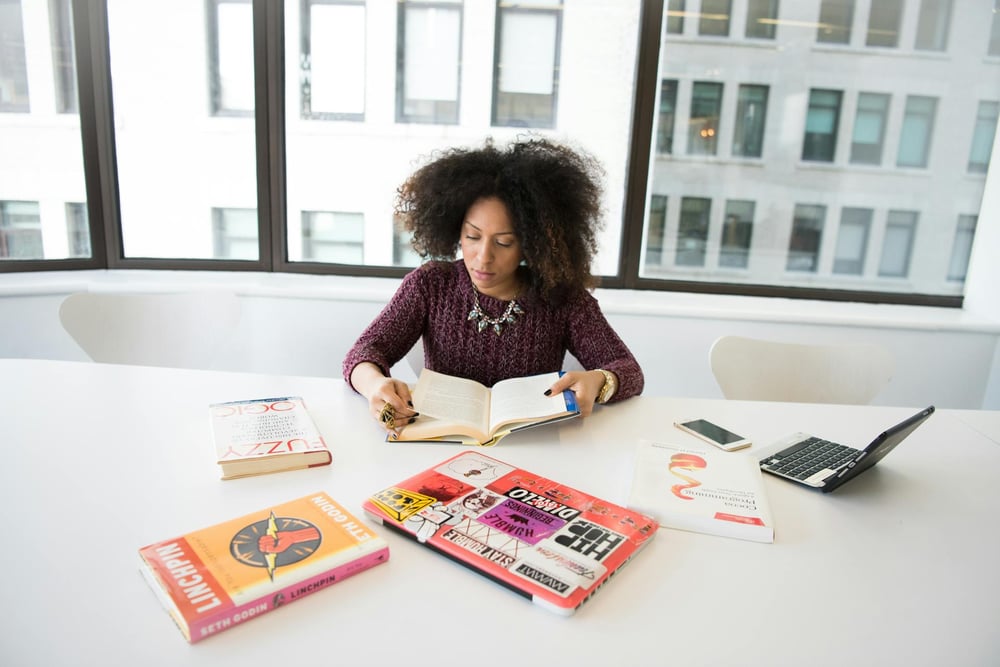 Several books and a couple laptops sit on a table. A person sits at the table reading a book.
