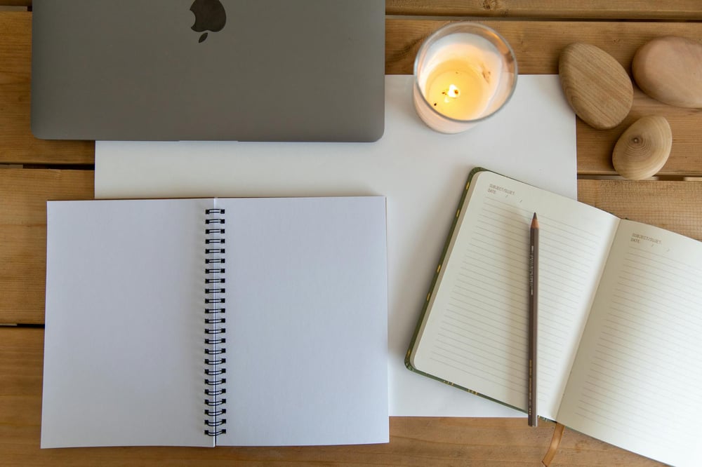 Two open notebooks and a laptop sit on a wooden table beside a lit candle and three rocks.