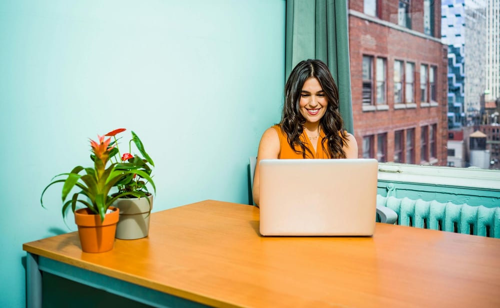 A person types on a laptop on a big wooden desk beside a window in a teal room.