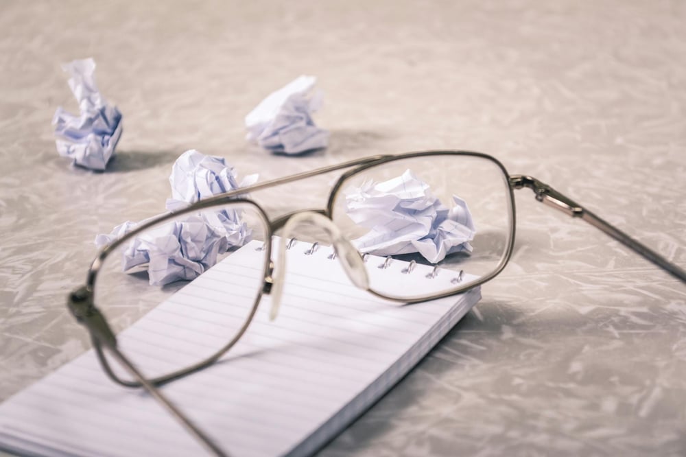 Glasses sit on top of a small notebook, surrounded by sheets of wadded up paper.