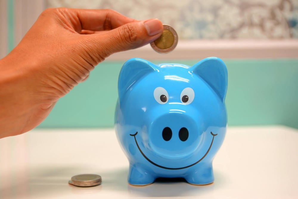 A hand drops a coin into a blue smiling piggy bank.
