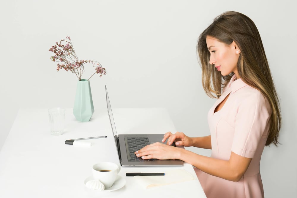 A person works on a laptop at a white desk near a vase of purple flowers.