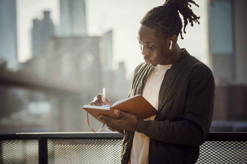 A person stands outdoors in a city, writing in a leatherbound journal.