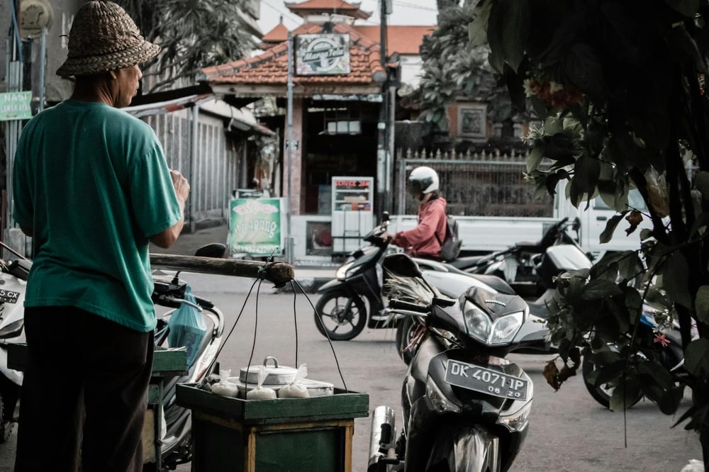 A person looks on as another person drives a motorbike down a street in Bali.