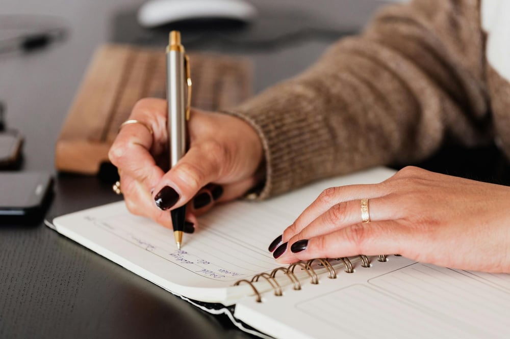 Hands wearing old rings and black nail polish write in a notebook.