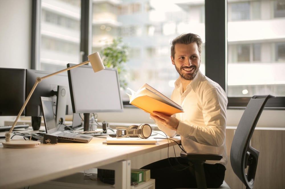 A person holding a book sits t a desk and smiles over their shoulder.