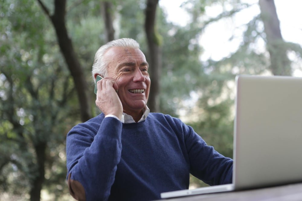 A smiling person talks on the phone while working on a laptop in a park.