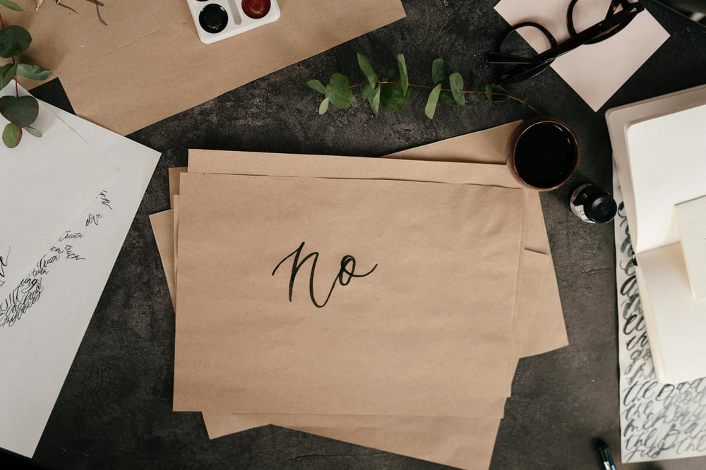 A stack of thin, brown sheets of paper sit on a desk alongside an inkwell, spectacles, and lettering projects. The word