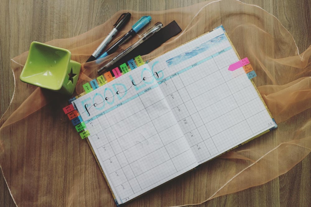 An open planner with several colorful tabs sits on a wooden table beside pens.