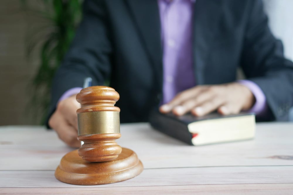 A person whose face is out of view bangs a gavel with their right hand and rests their left hand on a thick book.