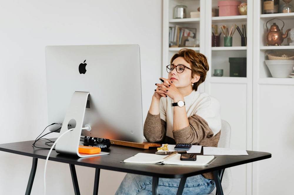 A person sits at a desk, folding their hands in front of their chin as they read the screen of a large desktop computer.