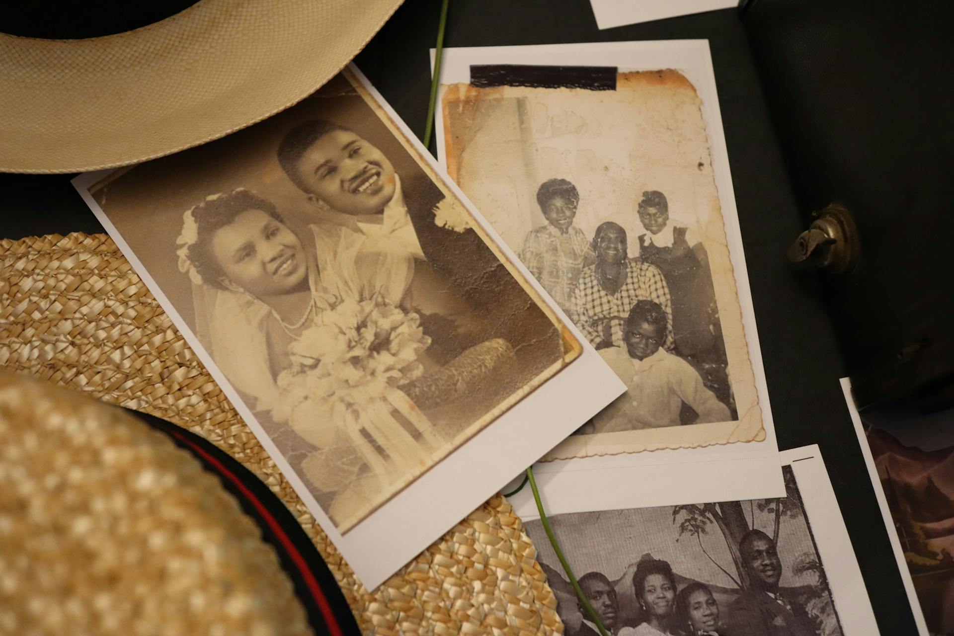 Black and white photographs including a wedding photo and family photo sit on a tabletop beside straw hats.