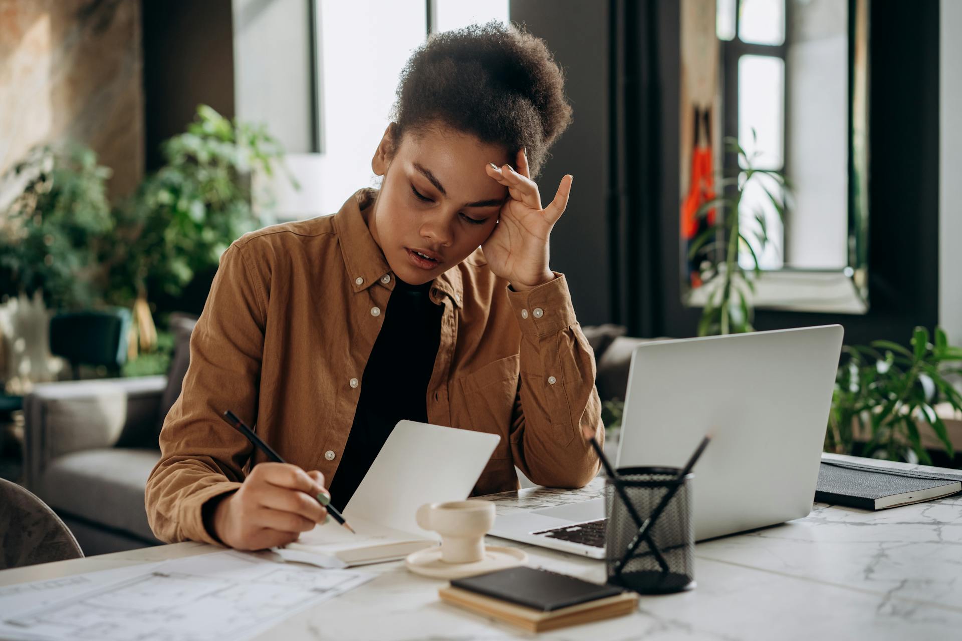 A person rests their head in one hand and writes in an notebook with the other. An open laptop sits in front of them on a desk.