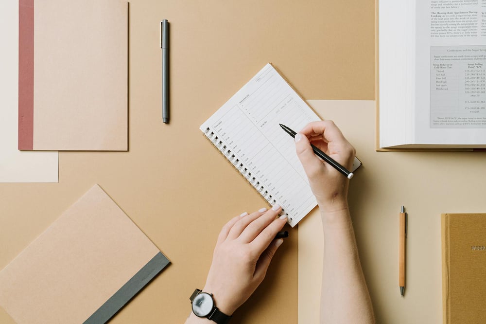 A hand writes in a planner. Notebooks, pens, and an open textbook surround the planner.
