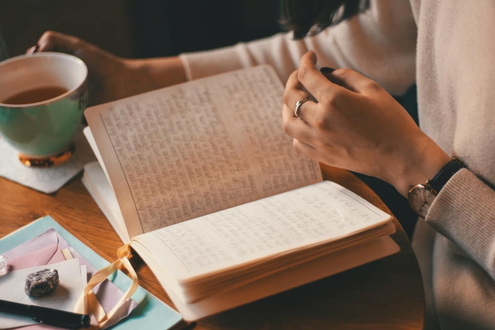 A person holds a cup of tea and looks at handwritten passages in a journal.
