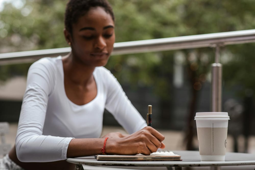 A person sits outside at a cafe table and writes in a notebook.