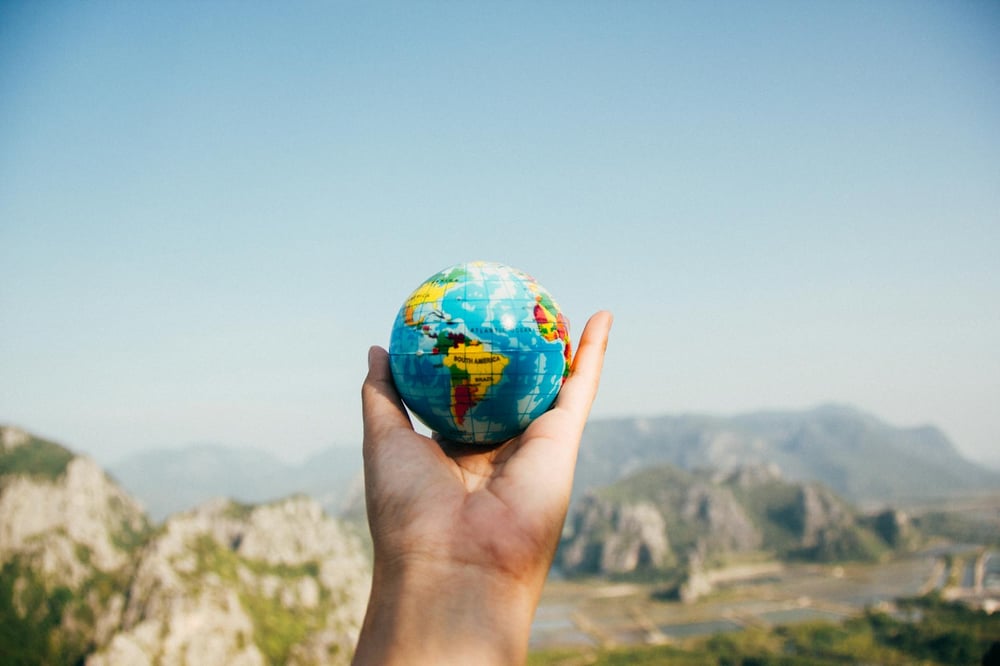 A hand holds out a small globe over a beautiful view of small mountains.