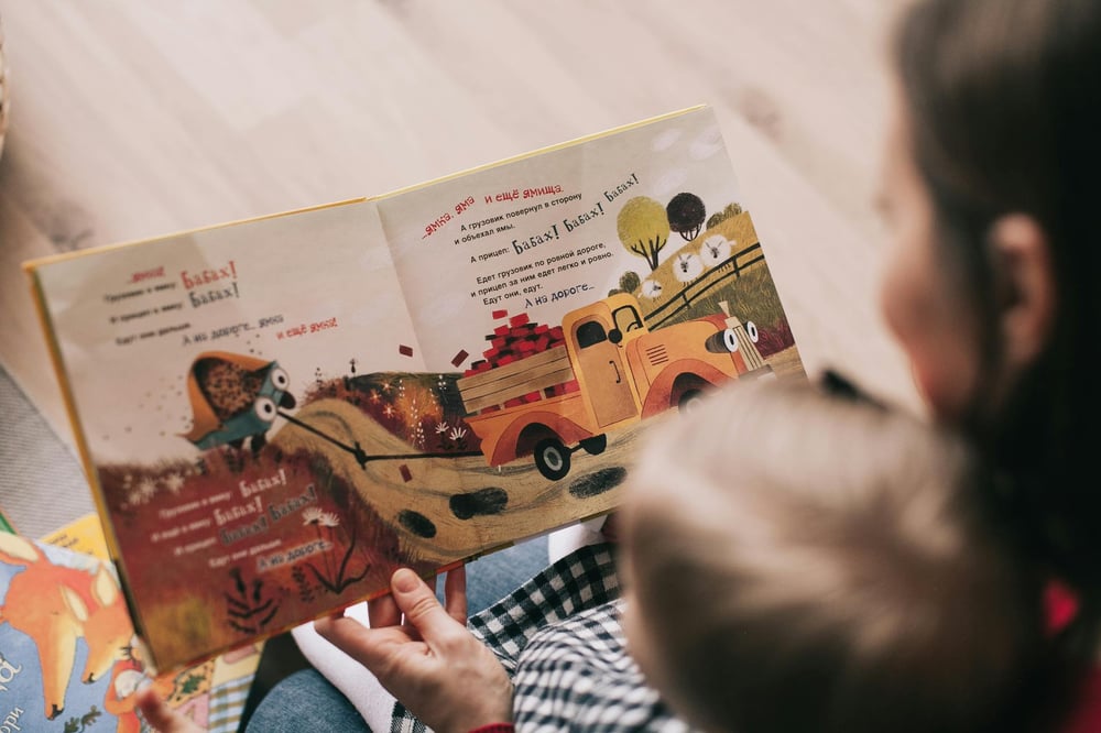 Over-the-shoulder view of a parent and small child reading a picture book in a non-English language.