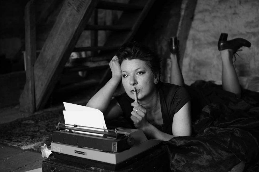 A person in a dress lies on the floor on their stomach in front of a typewriter, propped up on their elbows, holding a pen to their lips and thinking.