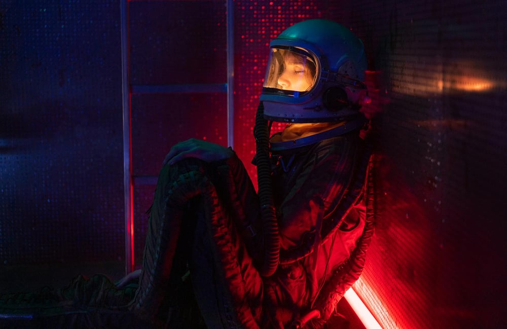 A person in a space suit sits on the floor of a dark room, lit only by a red light on the floor.