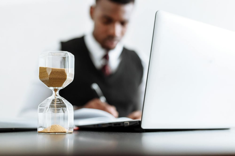 Sand moves through an hourglass as a blurred figure in the background sits in front of a laptop and writes in a notebook.