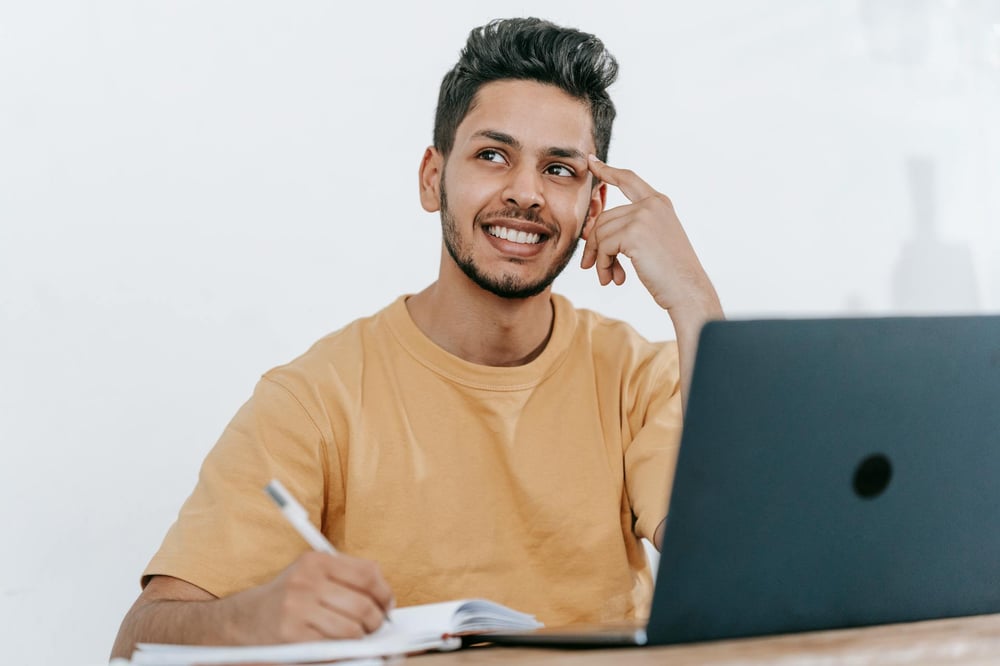 A person sitting at a laptop writes in a notebook with one hand and touches their other hand to their temple, staring off into space and thinking.t
