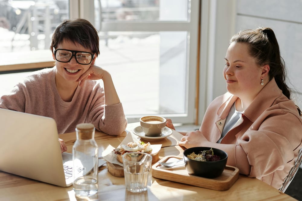 Two smiling people sit together in a cafe, looking at a laptop screen.