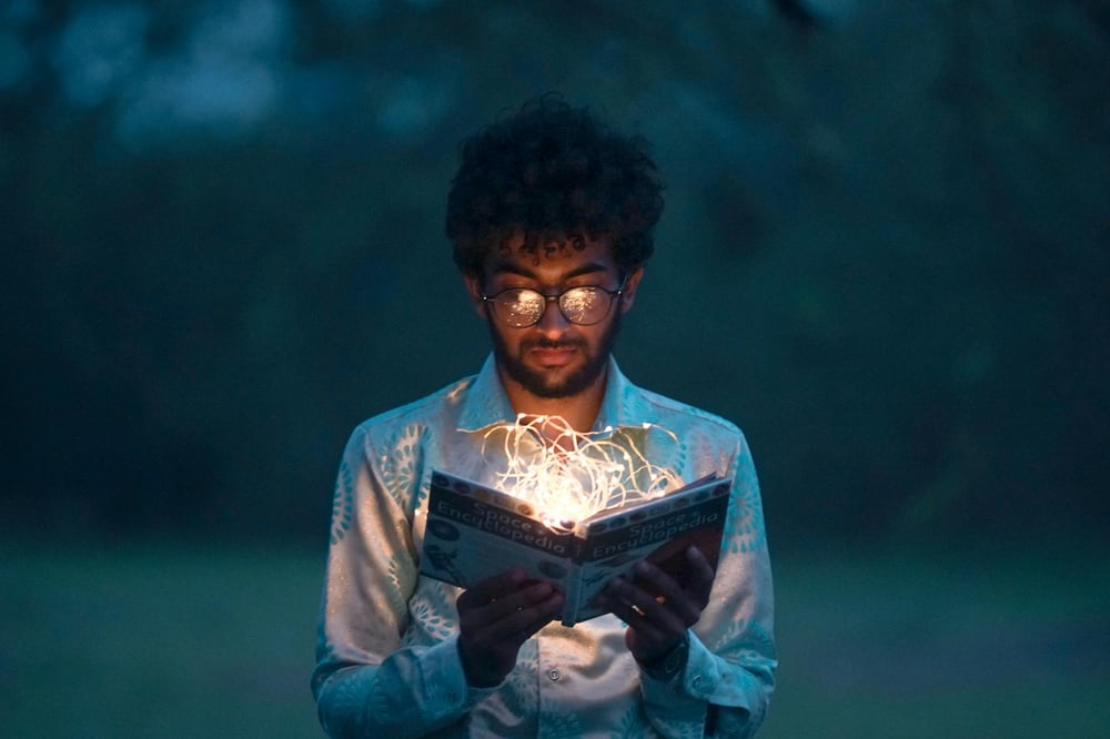 A person holds open a book with a tangle of string lights on the pages, illuminating the reader's face.