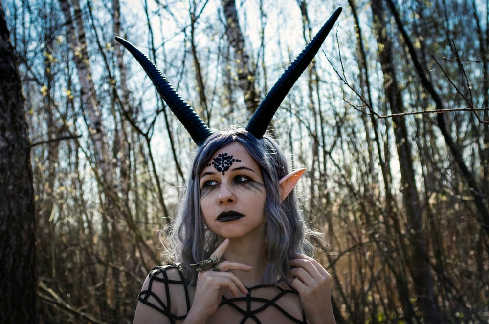 A fantasy character with pointy ears, long narrow horns, and a marking on their forehead touches a finger to their chin and looks off into the distance.