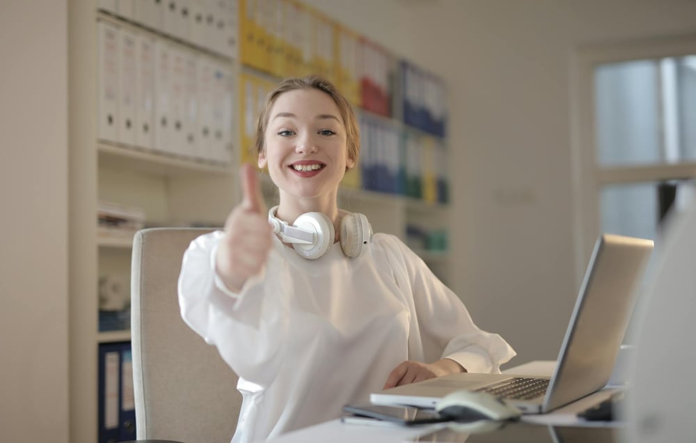 A person sitting at a computer and wearing headphones around their neck smiles and gives a thumbs up.