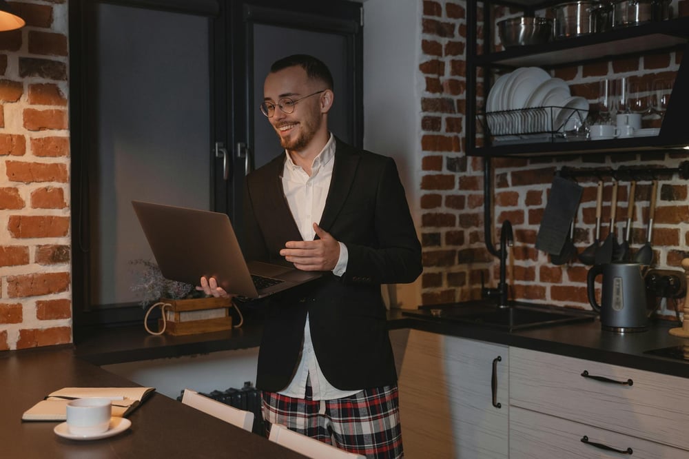 A person wearing a dress shirt, suit jacket, and pajama pants stands in a kitchen, taking a video meeting on a laptop.