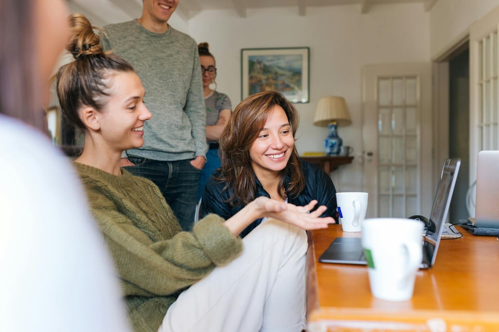 Five people gather around a laptop in a living room.