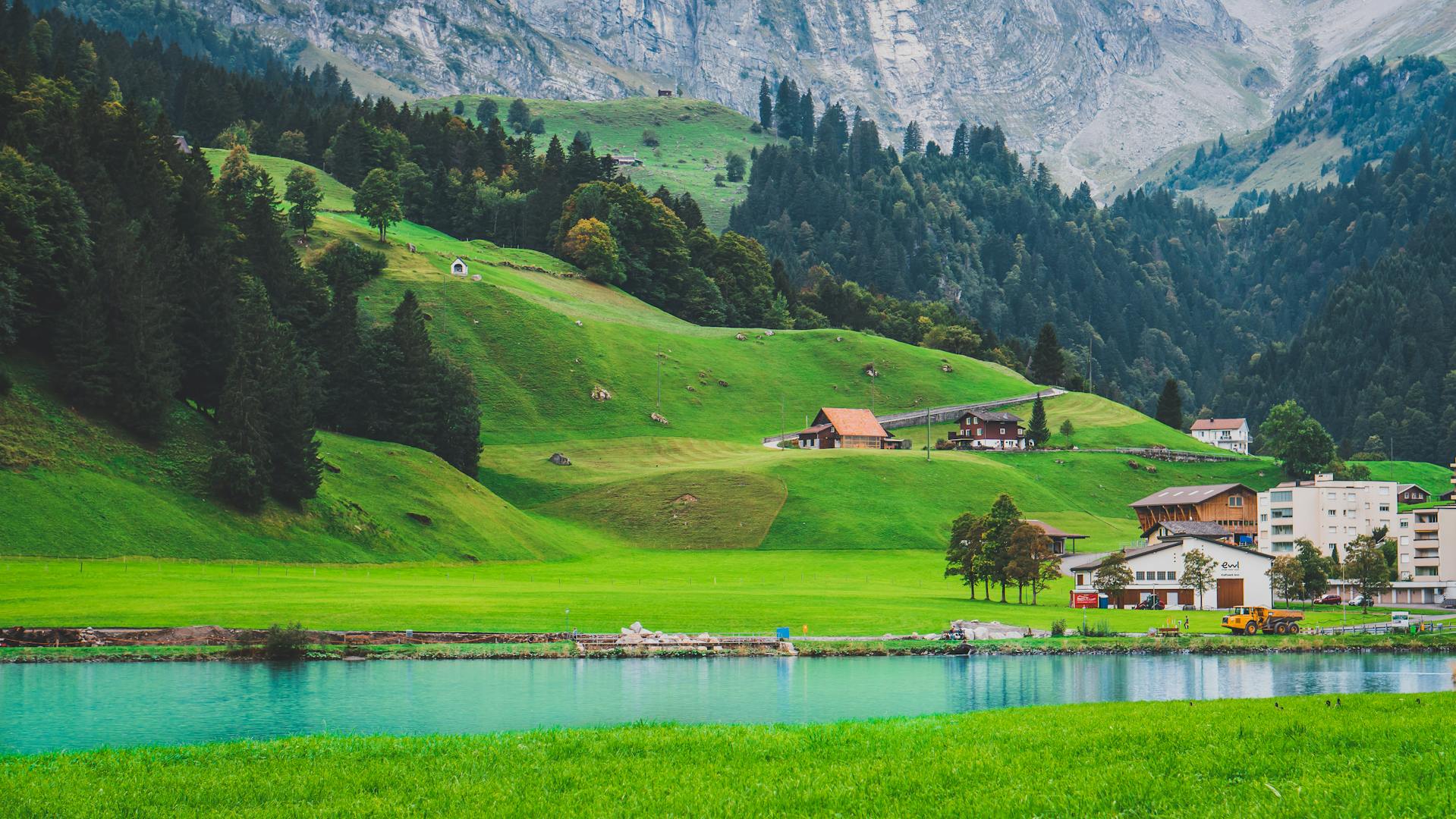 A beautiful scene of small buildings beside a blue lake on green hilly farmland backed by the stone wall of a mountain.