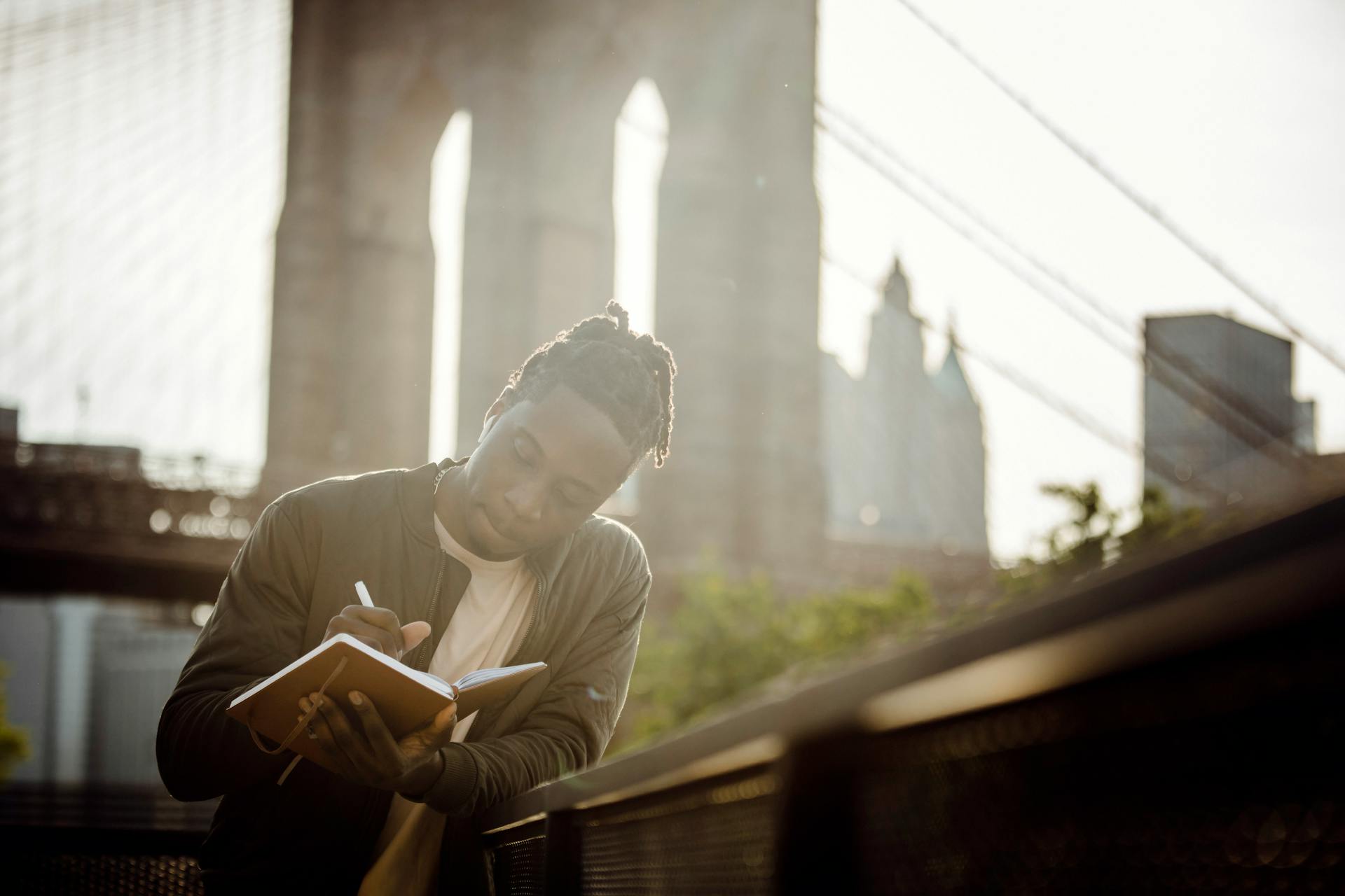A person stands outside, leaning against a wall and writing in a notebook. A bridge is in the background.