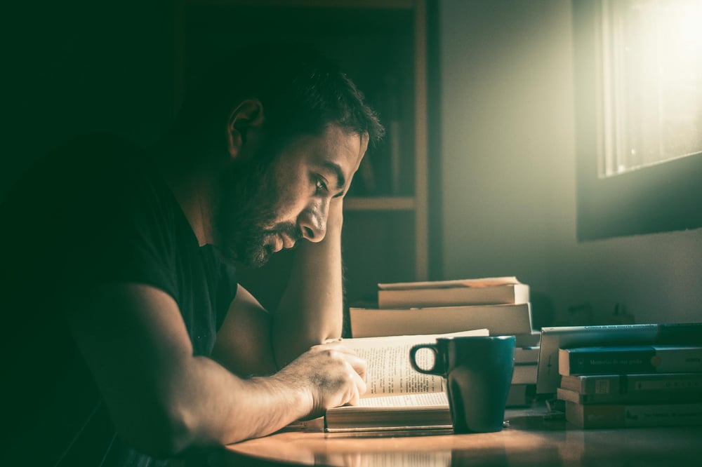 A person studies in the sunlight coming through the window of a dimly lit room.