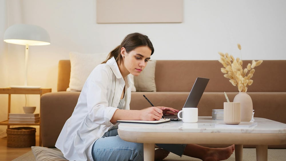 A writer sits on the floor in front of their coffee table, writing in a notebook and resting their free hand on the keyboard of an open laptop.