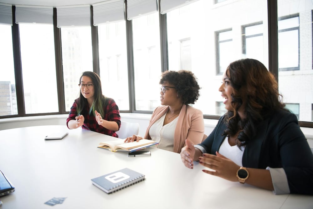 Three people sit at a conference room table, holding notebooks and talking animatedly.