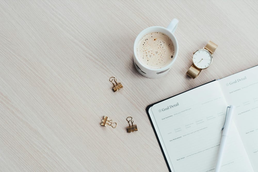 A project planning notebook lies open on a desk beside a cup of coffee, a watch, and three gold binder clips.
