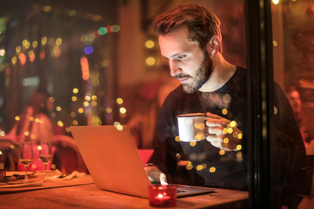 Behind a window reflecting streetlights, a writer sits in a café holding a cup of coffee and typing on a laptop.