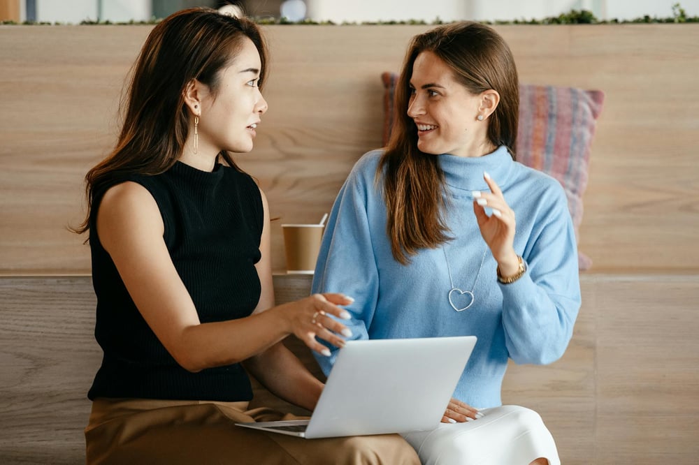 Two writers sit in front of a laptop, discussing something.