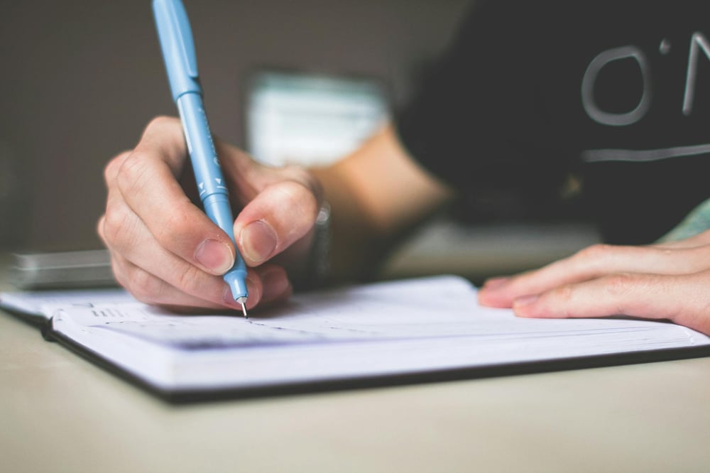 A hand holding a blue pen writes in a notebook.